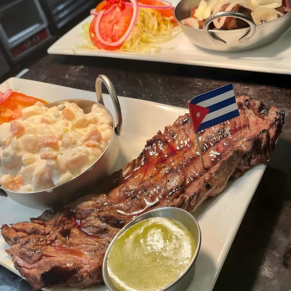 Grilled Cuban skirt steak with a Cuban flag, potato salad, and green sauce on a white plate.