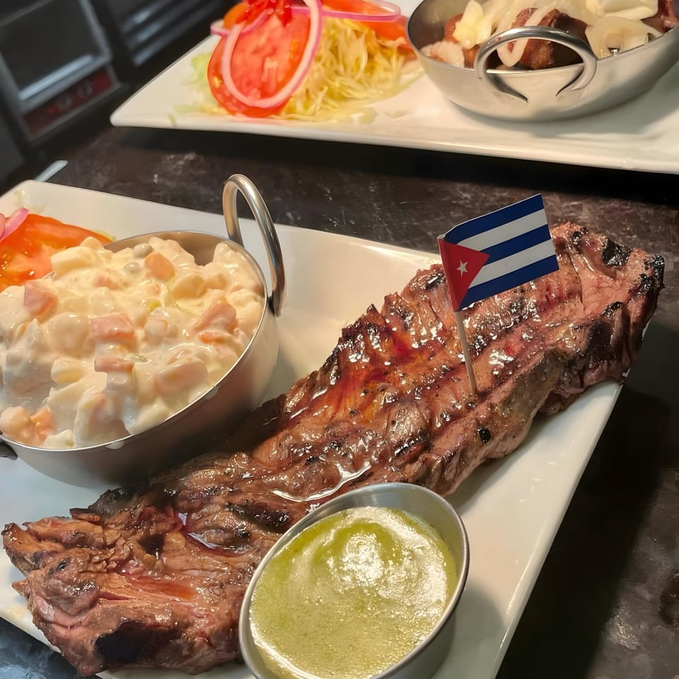 Grilled Cuban skirt steak with a Cuban flag, potato salad, and green sauce on a white plate.