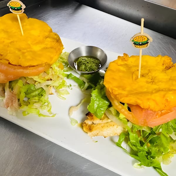 Two Cuban-style burgers with plantain buns, lettuce, tomato, and chicken, served with green sauce.