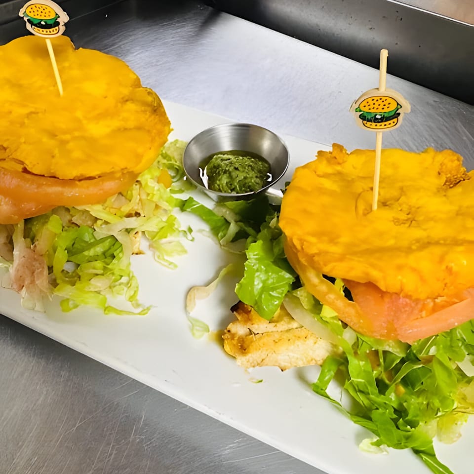 Two Cuban-style burgers with plantain buns, lettuce, tomato, and chicken, served with green sauce.