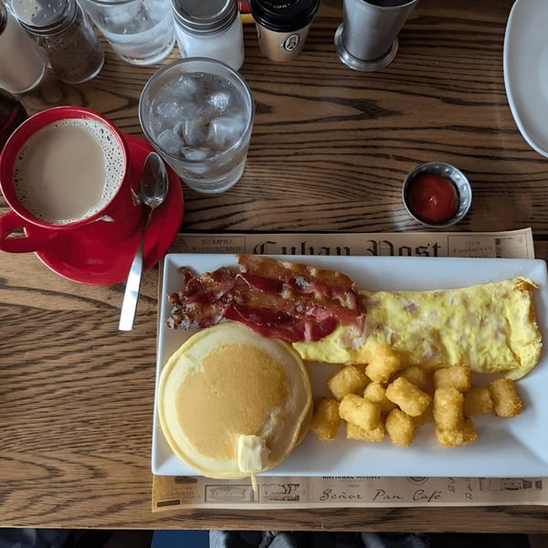 A Cuban breakfast with pancakes, bacon, omelet, tater tots, coffee, and water on a wooden table.