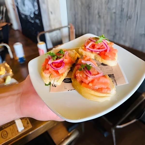 A hand holds a white plate with three Cuban bruschettas topped with tomatoes, red onions, and herbs.
